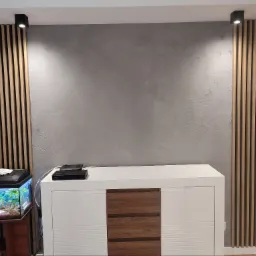 Szara ściana z efektem betonu, otoczona dekoracyjnymi panelami z drewnianych listewek, oświetlona punktowymi lampami sufitowymi. Poniżej biała komoda z drewnianymi szufladami i akwarium z roślinami...