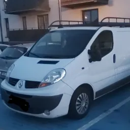 Biały Renault Trafic z relingami dachowymi zaparkowany na parkingu, widoczne inne samochody i budynek mieszkalny w tle.