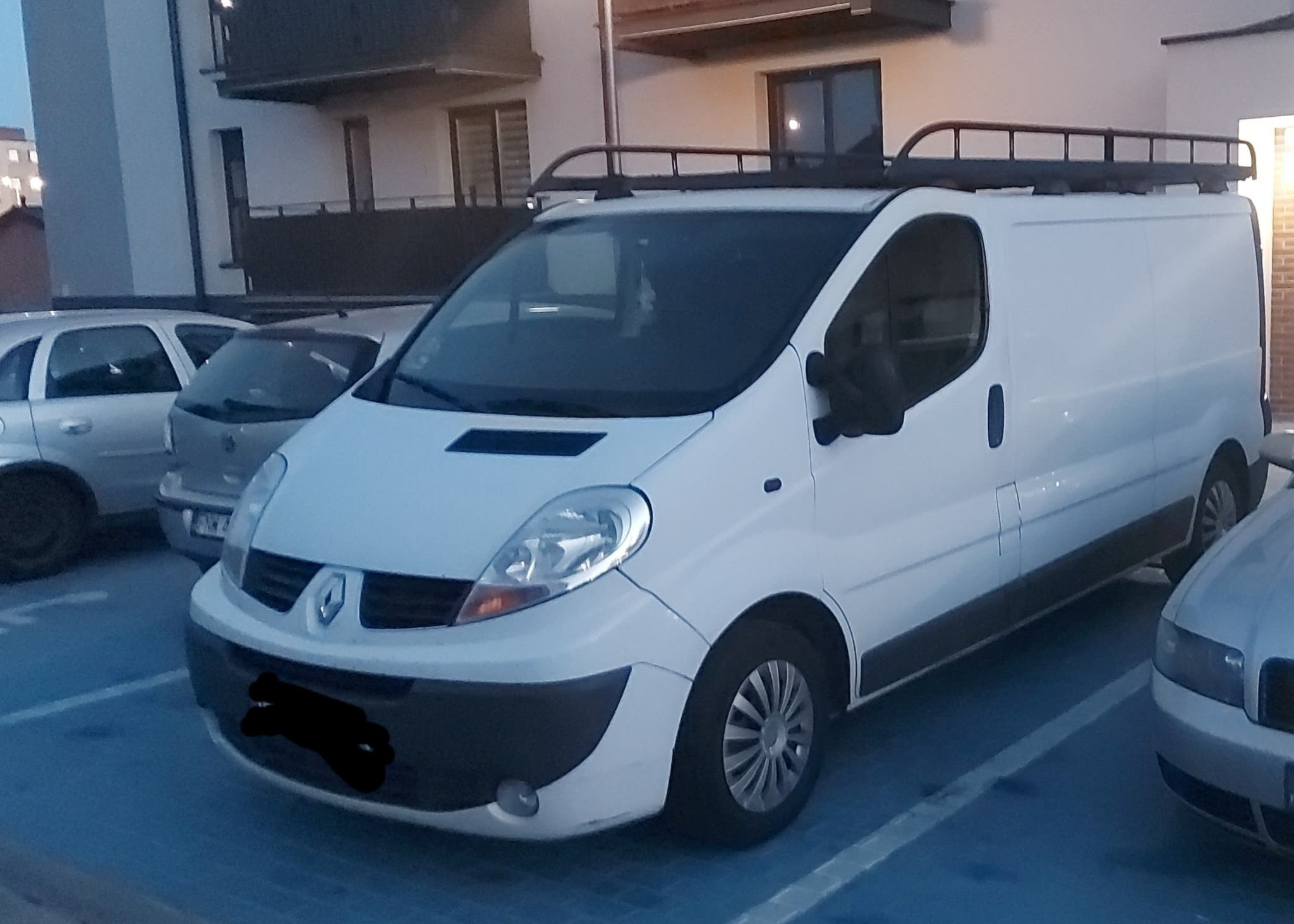 Biały Renault Trafic z relingami dachowymi zaparkowany na parkingu, widoczne inne samochody i budynek mieszkalny w tle.