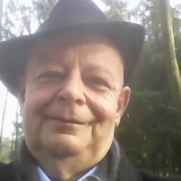 Portret mężczyzny w kapeluszu na tle drzew, uśmiechający się do kamery.