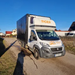 Biały samochód dostawczy z czarną plandeką i pomarańczowym napisem TIGER na tle błękitnego nieba, cień fotografa na drodze.