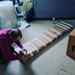 Mężczyzna w fioletowej bluzie klęczy na podłodze, pakując długi, płaski przedmiot w kartonowe pudełko i zabezpieczając go białą taśmą klejącą.