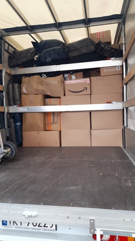 Wnętrze samochodu dostawczego wypełnione kartonami i workami, widoczne oznaczenia 'FRAGILE' i 'GLASS' na niektórych paczkach, na podłodze widoczny numer rejestracyjny pojazdu.