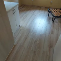 Jasne panele podłogowe z widoczną strukturą drewna, ułożone w pokoju z meblami i oknem w tle, widoczne listwy przypodłogowe.