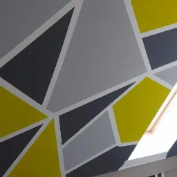 Ściana pod skosem na poddaszu pomalowana w geometryczne wzory z użyciem szarego, grafitowego i żółtego koloru, oddzielone białymi liniami, z oknem dachowym w tle.