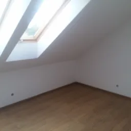 Poddasze z oknem dachowym i drewnianą podłogą, świeżo po malowaniu ścian na biało, widoczne dwa otwory w ścianie na instalacje.