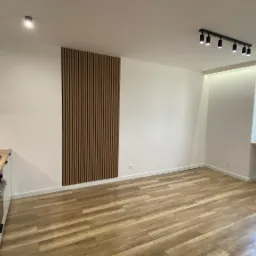 Nowoczesne wnętrze mieszkania po remoncie: drewnopodobna podłoga, białe ściany z dekoracyjnym panelem pionowym, aneks kuchenny z czarną armaturą i oświetleniem punktowym.