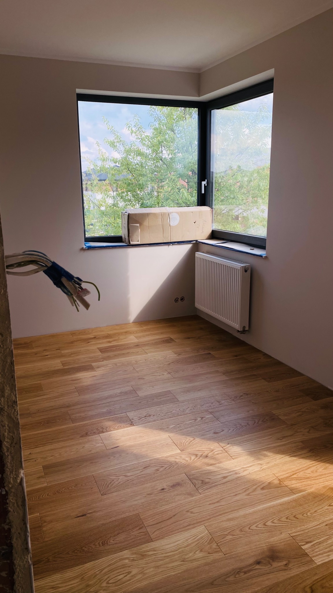 Pokój po remoncie z nową drewnianą podłogą, grzejnikiem i dużym narożnym oknem z widokiem na zieleń; wiązka kabli wystaje ze ściany po lewej stronie.