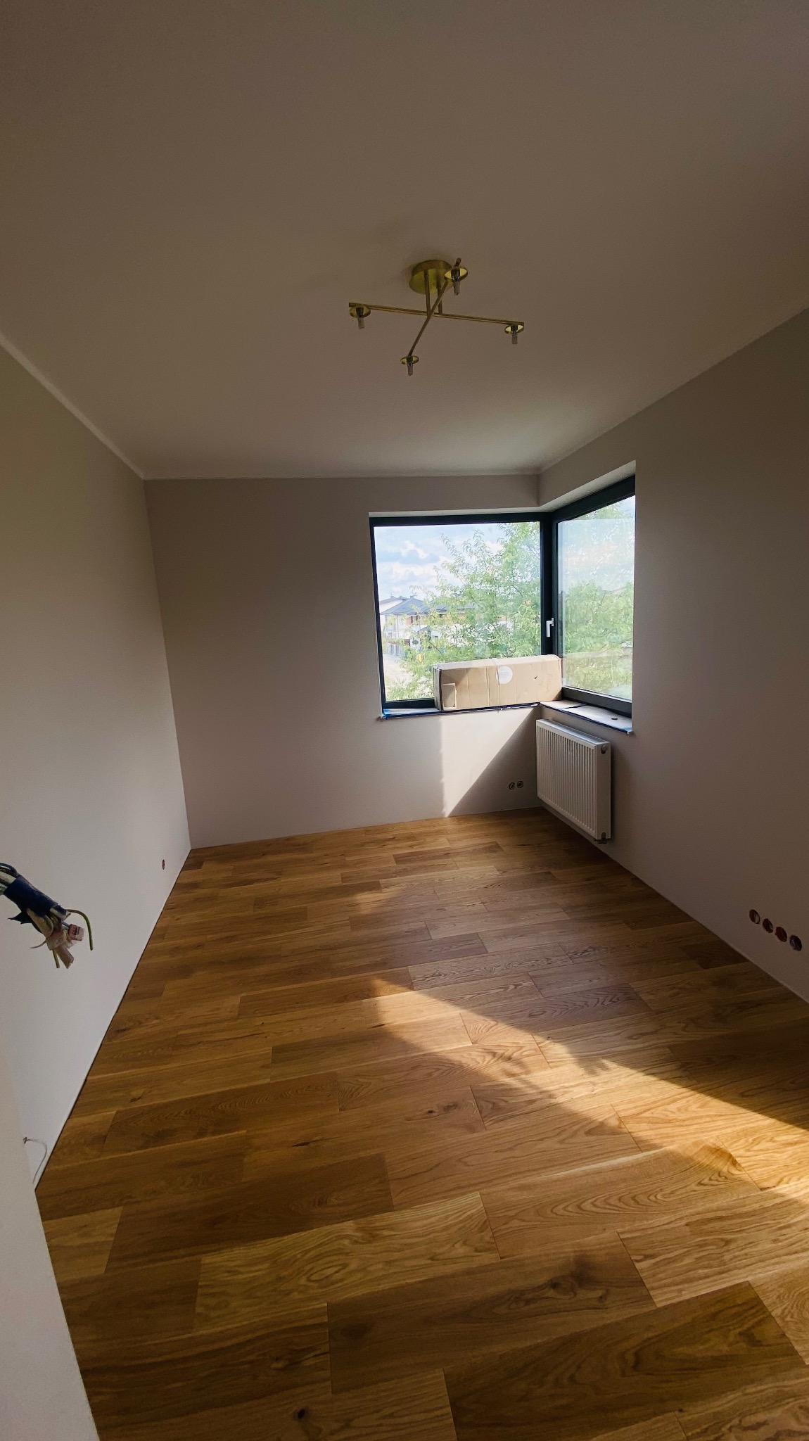 Pokój po remoncie z drewnianą podłogą, szarymi ścianami i oknem z widokiem na zieleń; na parapecie kartonowe opakowanie.