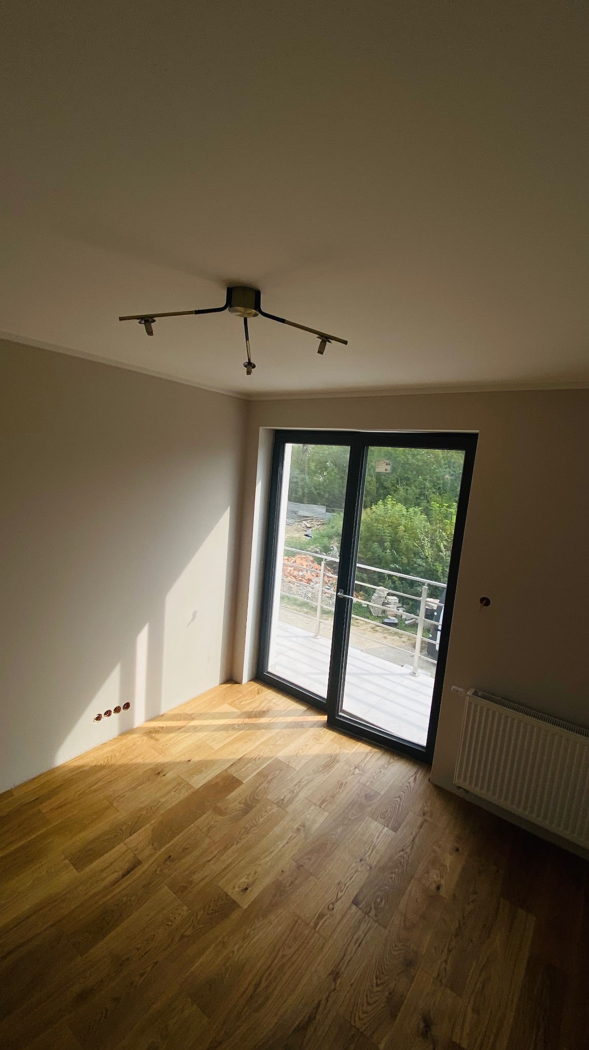 Jasne, puste pomieszczenie z parkietem, świeżo pomalowanymi ścianami i dużym oknem balkonowym wychodzącym na zieleń, widoczne gniazdka elektryczne w ścianie i nowoczesna lampa sufitowa.