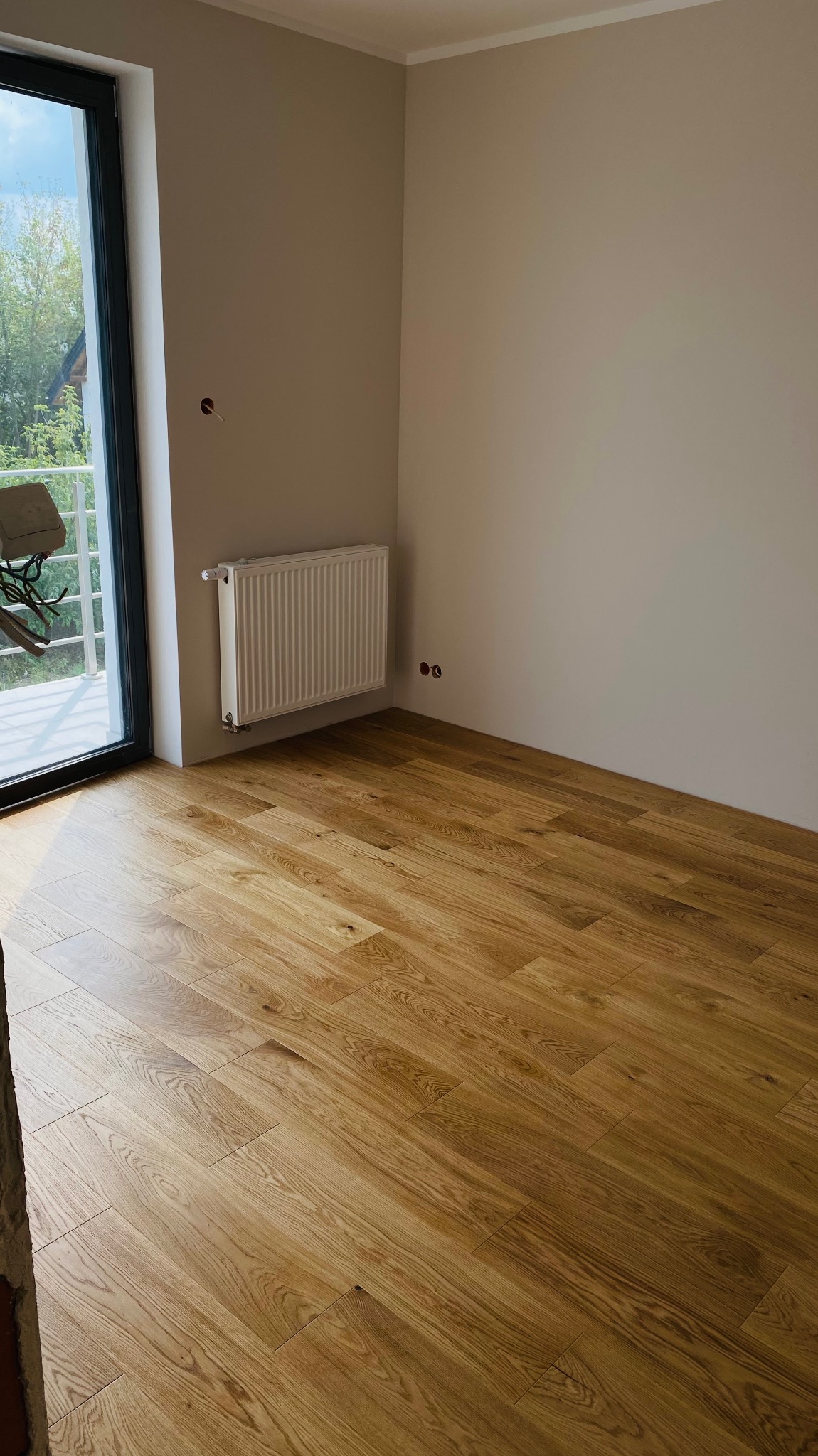 Jasne panele podłogowe ułożone w nowym pokoju z grzejnikiem pod ścianą i dużym oknem balkonowym.