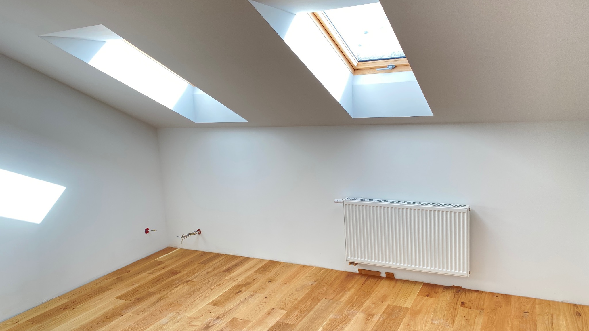 Poddasze po remoncie z drewnianą podłogą, białymi ścianami, grzejnikiem i dwoma oknami dachowymi, widoczne niedokończone instalacje elektryczne.