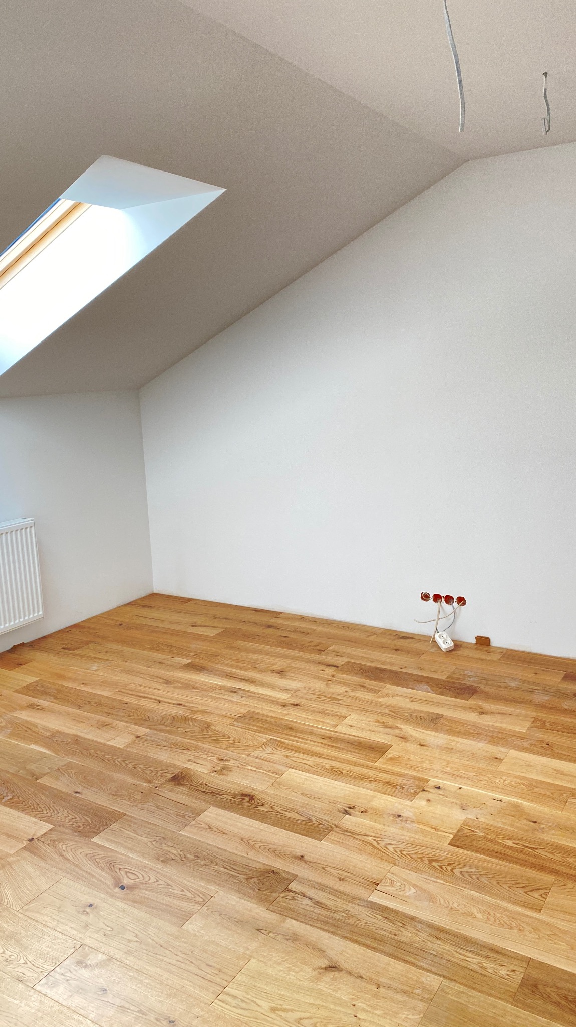 Poddasze po remoncie: jasne drewniane panele podłogowe, białe ściany z oknem dachowym, widoczne gniazdka elektryczne bez osprzętu.