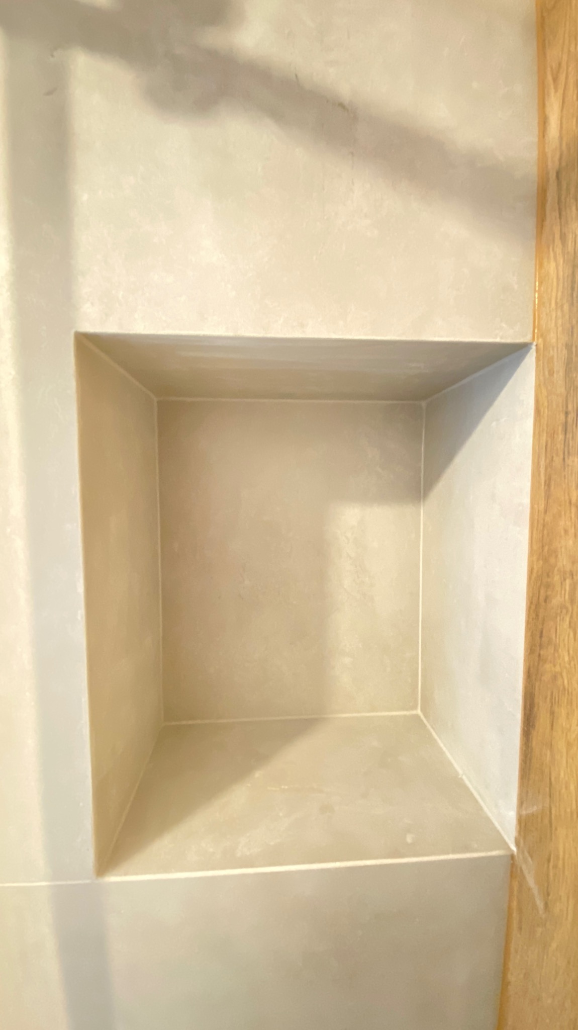 Wnęka w ścianie łazienki, wykończona jasną, beżową gładzią, z widoczną fakturą i delikatnym połyskiem, obok fragment drewnianej boazerii.