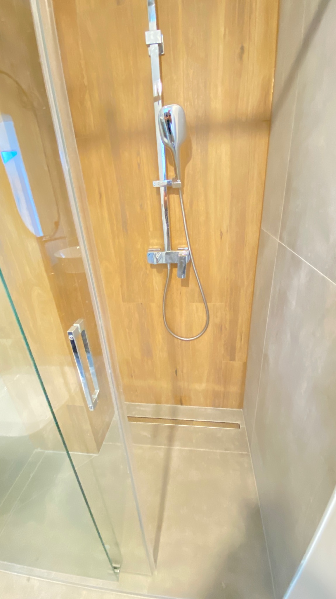 Nowoczesna kabina prysznicowa z drewnopodobną ścianą, chromowaną armaturą i odpływem liniowym w posadzce.