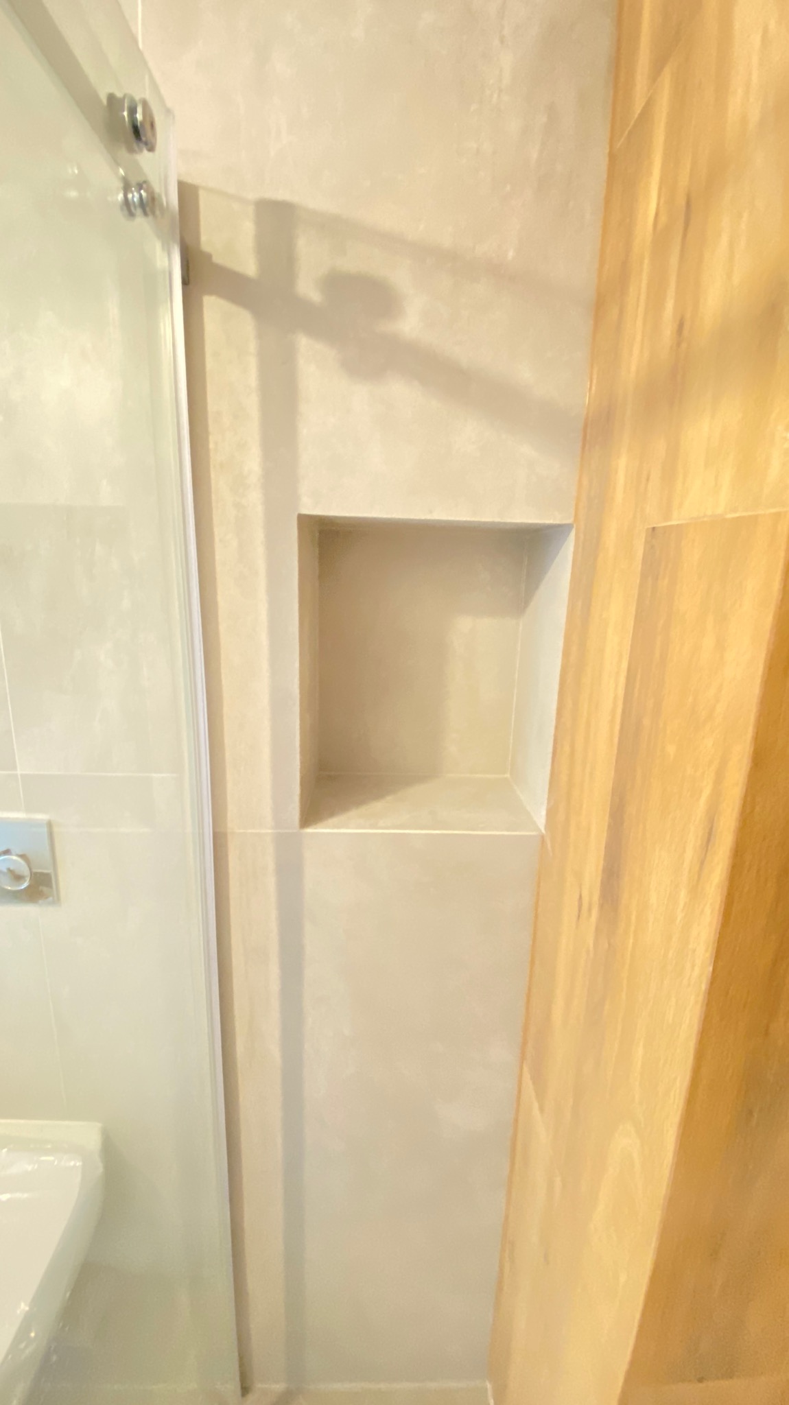 Beżowa łazienka z wnęką w ścianie wykończoną płytkami, widoczny fragment kabiny prysznicowej i drewniana listwa.