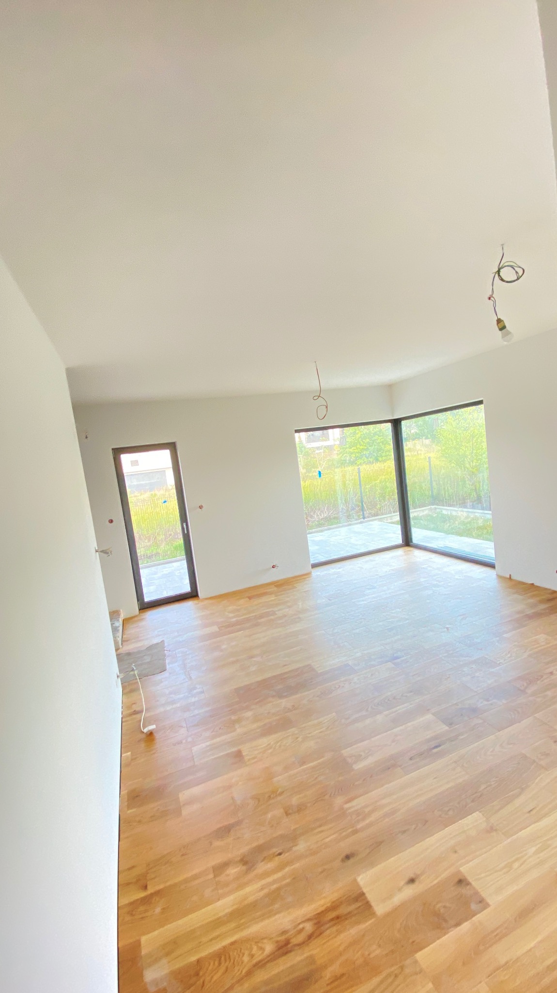 Jasne, przestronne wnętrze z parkietem w odcieniach dębu, białymi ścianami i dużymi oknami z widokiem na zieleń, z widocznymi przewodami oświetleniowymi wystającymi z sufitu.