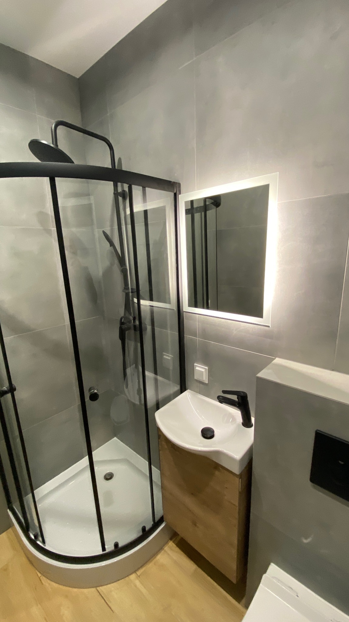 Nowoczesna łazienka z kabiną prysznicową z czarnymi profilami, umywalką nablatową na drewnianej szafce i podświetlanym lustrem.