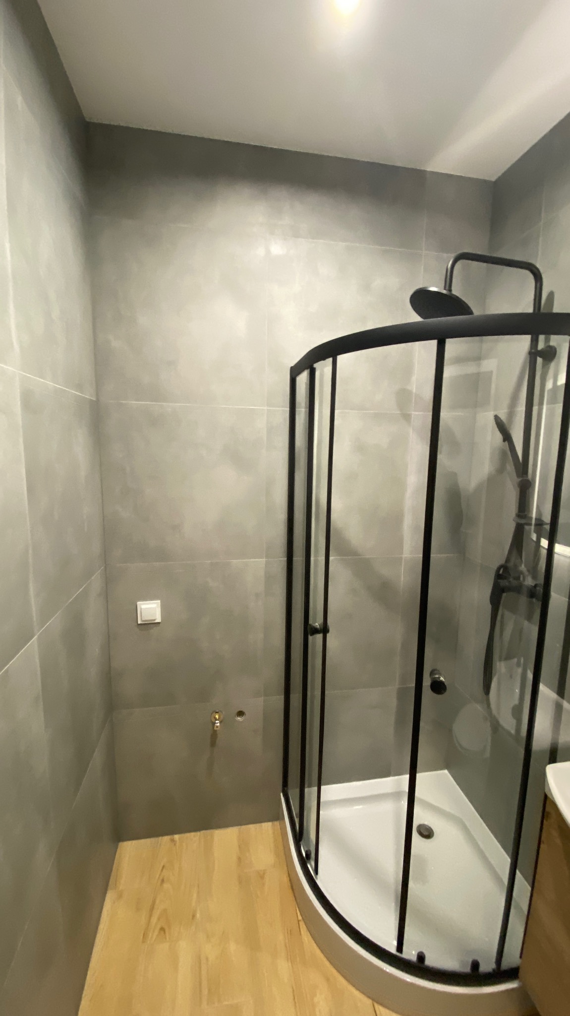 Nowoczesna kabina prysznicowa z czarnymi profilami i szarymi płytkami w łazience, widoczny fragment drewnopodobnej podłogi i przygotowane przyłącze wodne.