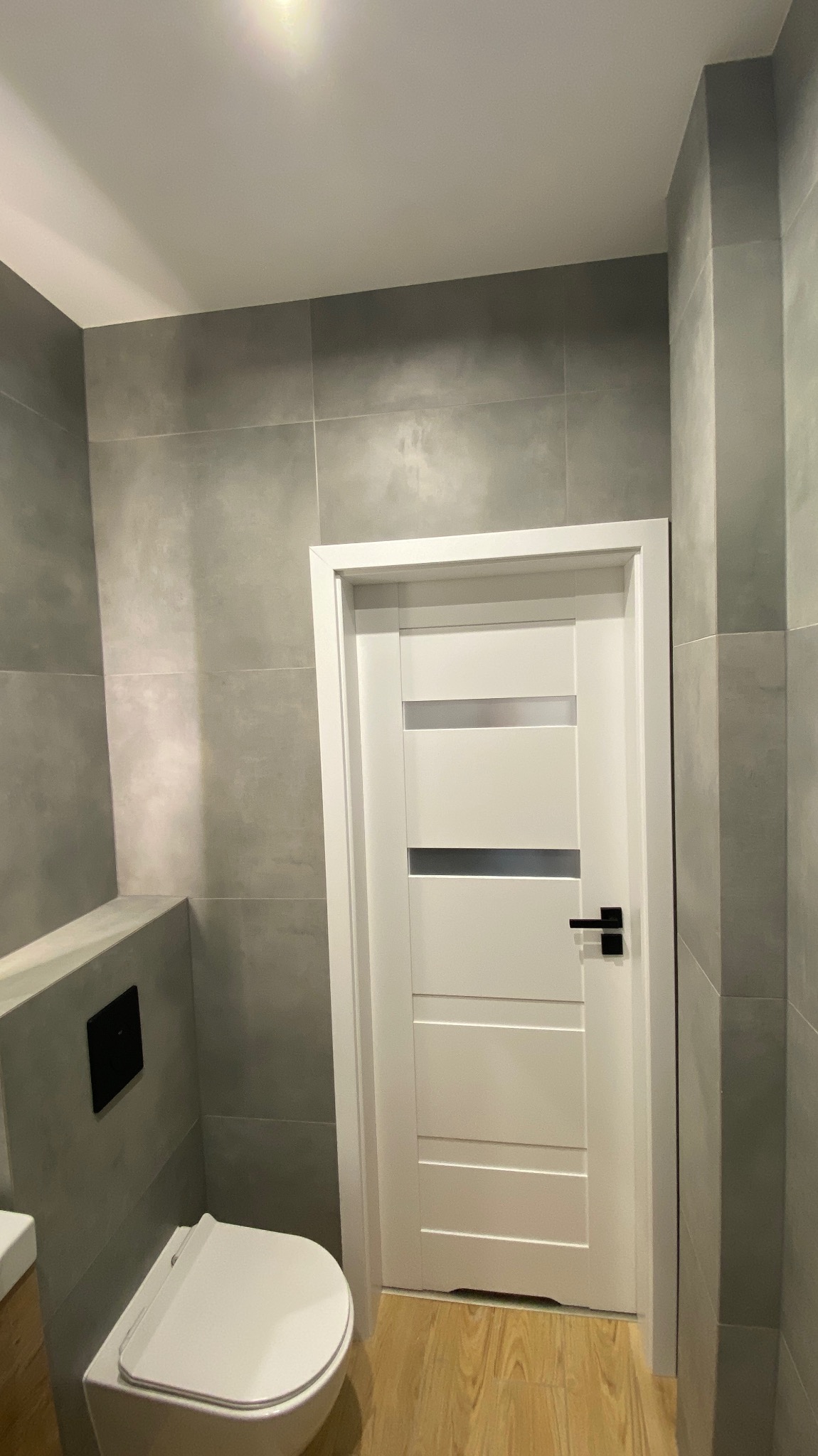 Nowoczesna łazienka z szarymi płytkami, podwieszana toaleta i białe drzwi z czarną klamką, widok z góry na świeżo wykończone wnętrze.