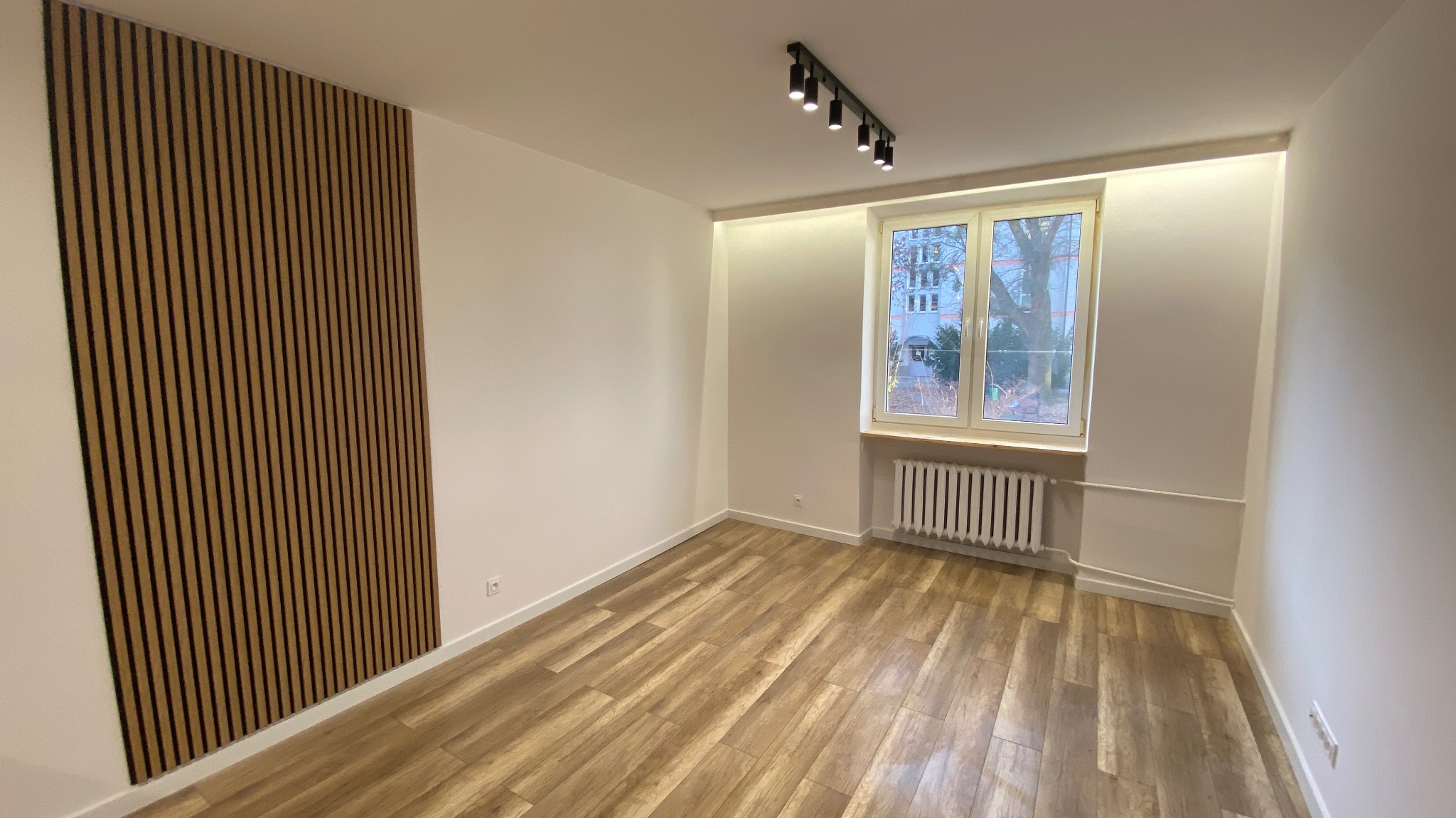 Pusty pokój po remoncie z drewnianą okładziną ścienną, panelami podłogowymi imitującymi drewno i nowoczesnym oświetleniem sufitowym w postaci czarnej listwy z reflektorami.