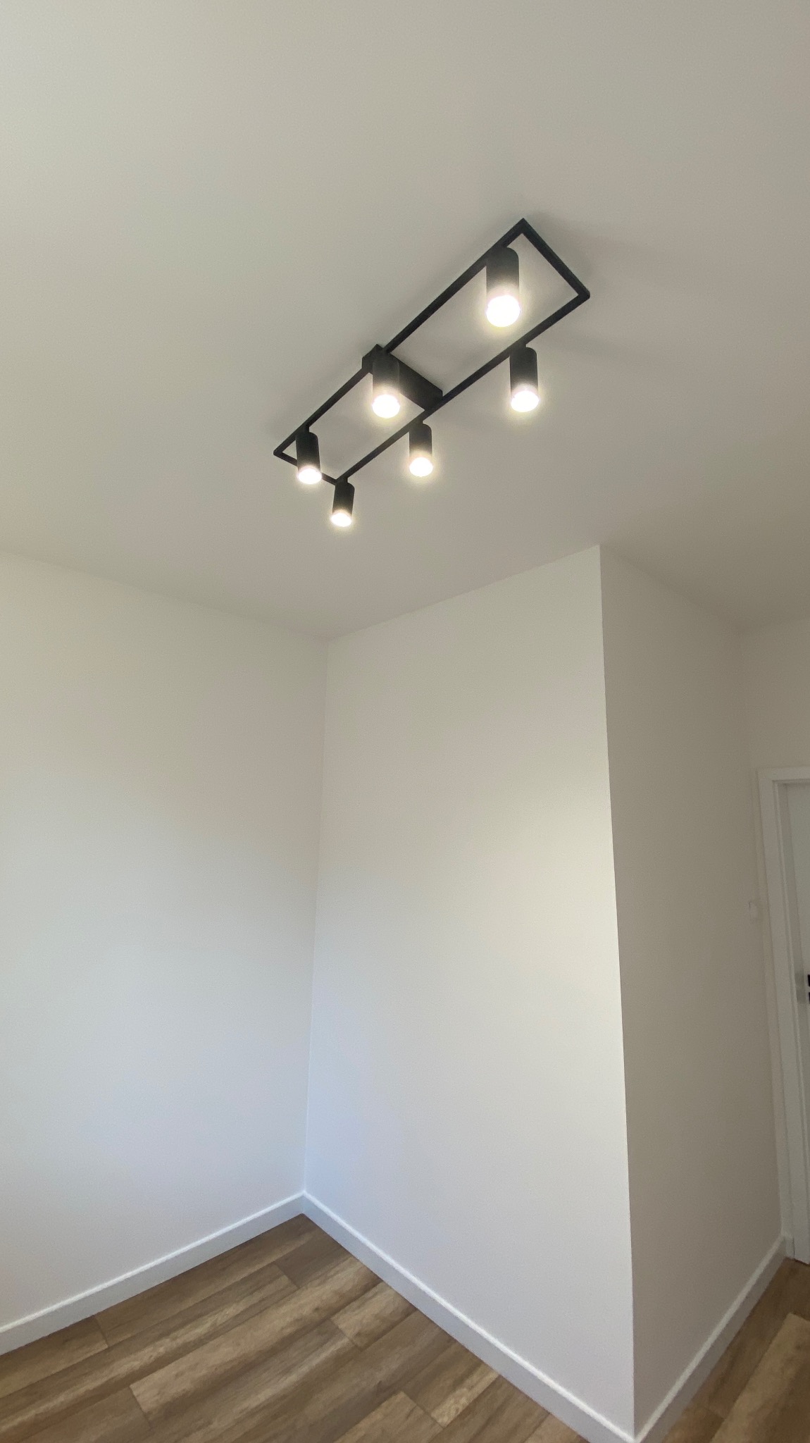 Nowoczesne oświetlenie sufitowe z sześcioma reflektorami w czarnej, prostokątnej oprawie w świeżo pomalowanym na biało pomieszczeniu z panelami podłogowymi.