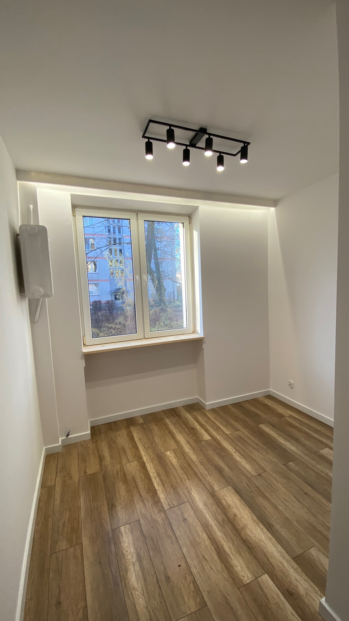 Pusty pokój po remoncie z drewnopodobną podłogą, białymi ścianami, oknem z widokiem na budynek i czarną lampą sufitową z sześcioma reflektorami.