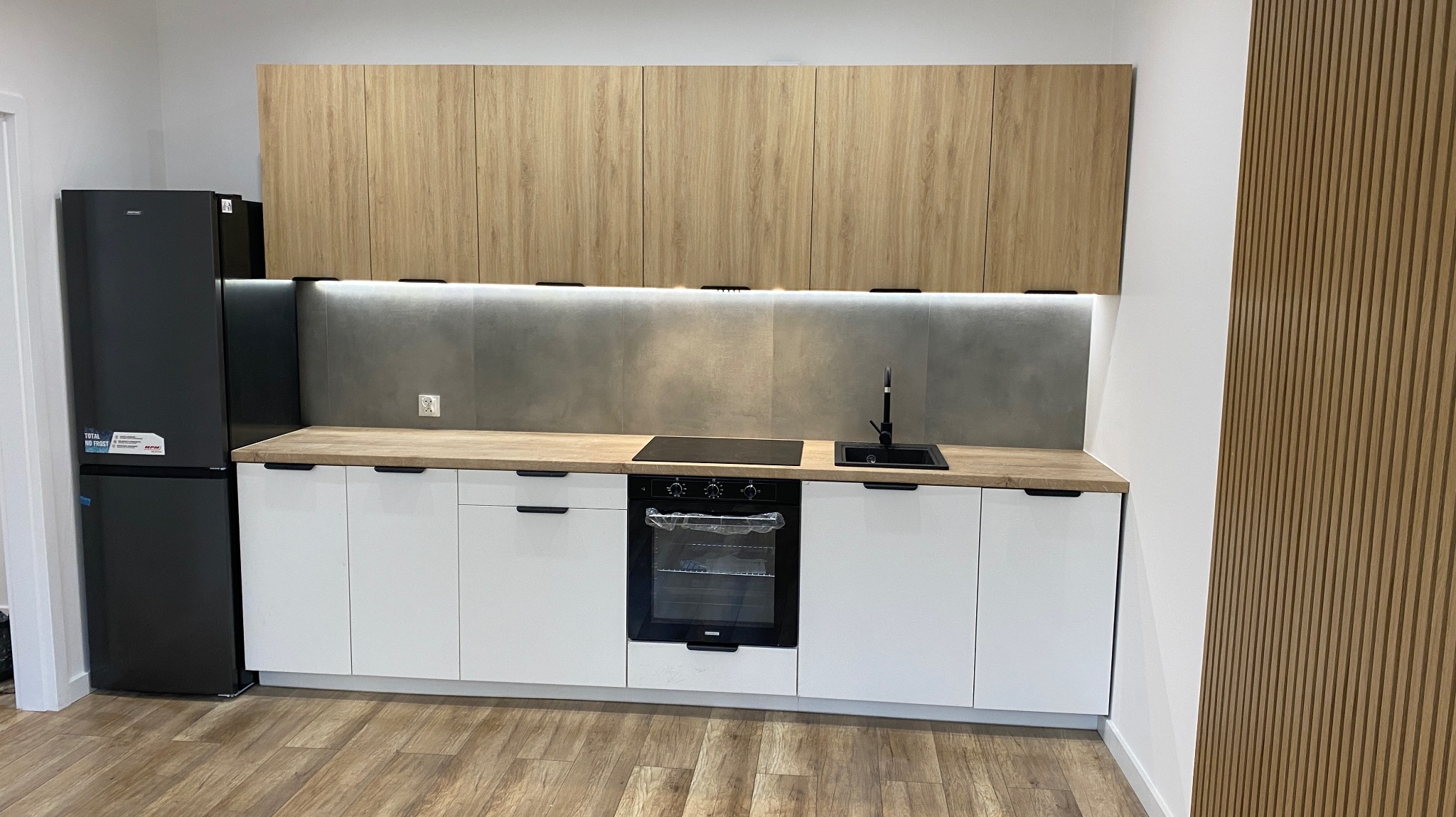 Nowoczesna kuchnia z białymi szafkami dolnymi i drewnopodobnymi górnymi, szarym panelem nad blatem, czarną lodówką i drewnianymi lamelami na ścianie po prawej.