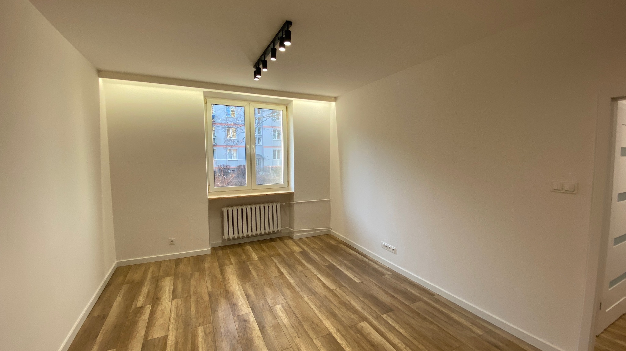 Pusty pokój po remoncie z drewnopodobną podłogą, białymi ścianami, oknem z widokiem na budynek i czarną listwą oświetleniową na suficie.