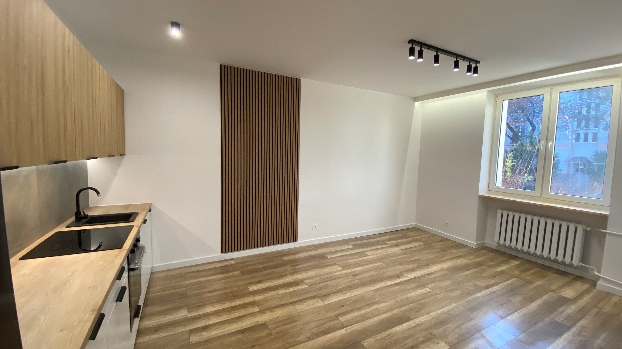 Nowoczesne wnętrze mieszkania po remoncie: drewnopodobna podłoga, białe ściany z dekoracyjnym panelem pionowym, aneks kuchenny z czarną armaturą i oświetleniem punktowym.