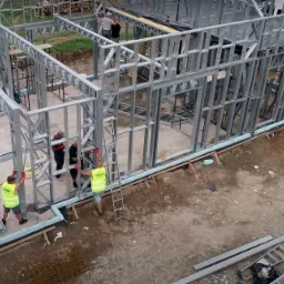 Montaż stalowej konstrukcji domu, widok z góry, pracownicy w kamizelkach odblaskowych, elementy konstrukcyjne na ziemi.