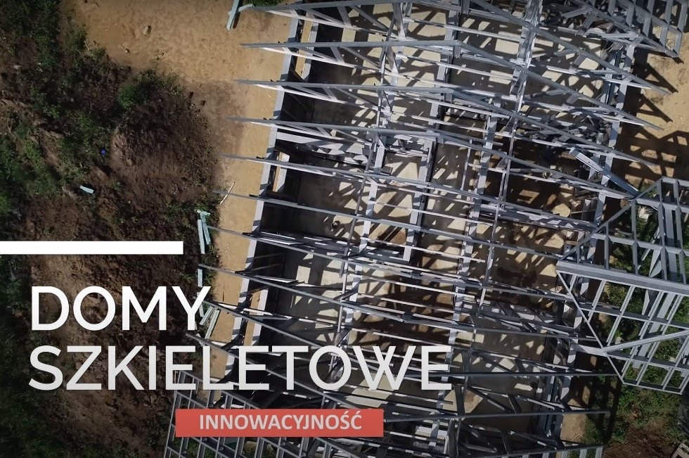 Widok z góry na metalowy szkielet domu w trakcie budowy, widoczne elementy konstrukcyjne i teren budowy.