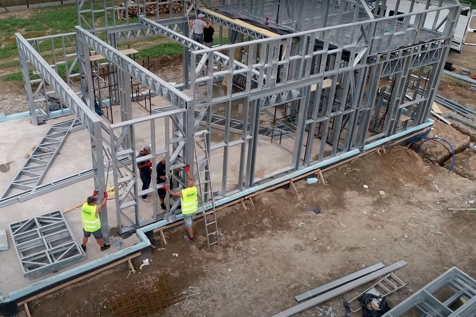 Montaż stalowej konstrukcji domu, widok z góry, pracownicy w kamizelkach odblaskowych, elementy konstrukcyjne na ziemi.