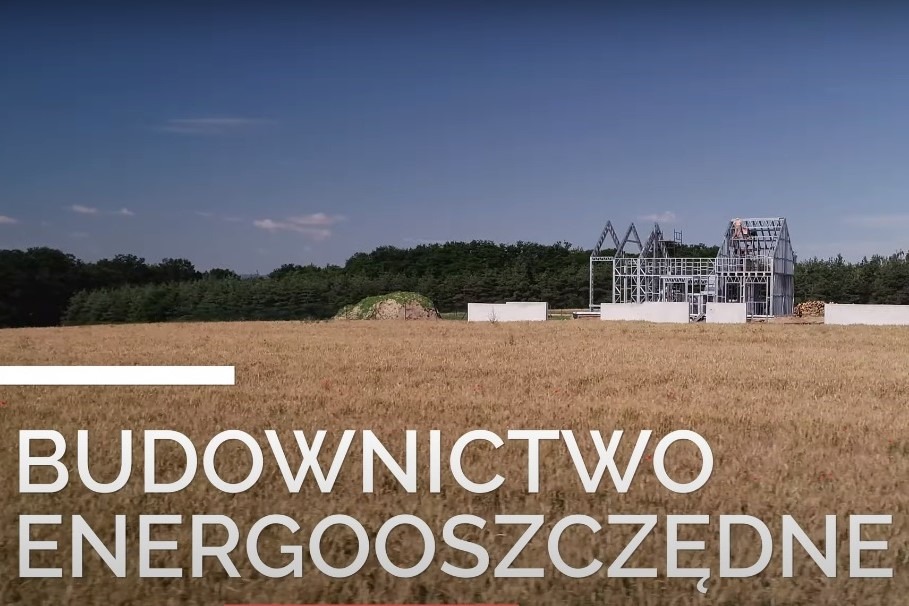 Wznoszenie konstrukcji domu szkieletowego na tle pola i lasu, widoczne białe płyty fundamentowe i elementy stalowe.