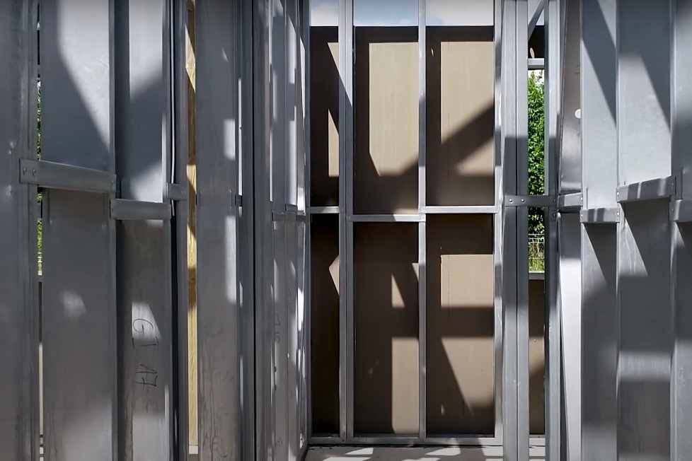 Konstrukcja szkieletowa domu w trakcie budowy, widoczne metalowe profile, płyty OSB i fragment zieleni w tle.