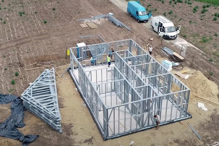 Budowa domu ze stalowym szkieletem: widok z góry na fundament i wznoszone ściany, z pracownikami wewnątrz konstrukcji i dwoma zaparkowanymi busami obok.
