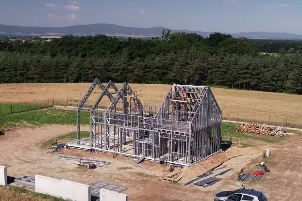 Konstrukcja szkieletowa domu jednorodzinnego w trakcie montażu, widoczni dwaj pracownicy na dachu, w tle krajobraz z polami i lasem.