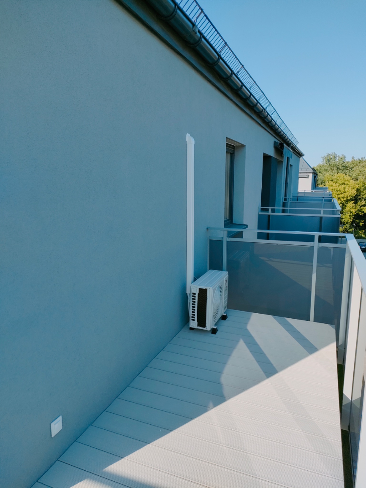 Zewnętrzna jednostka klimatyzacji zamontowana na balkonie budynku wielorodzinnego z szarą elewacją i drewnianą podłogą, widoczny biały przewód odprowadzający wodę, jasne światło słoneczne.