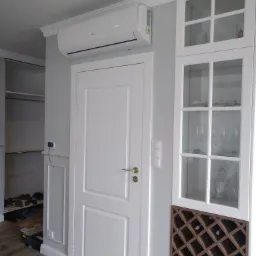 Biała jednostka klimatyzacyjna zamontowana nad białą, dwuskrzydłową wewnętrzną drzwią w pomieszczeniu z białą zabudową meblową z przeszklonymi witrynami i regałem na wino, widoczne narzędzia i buty...
