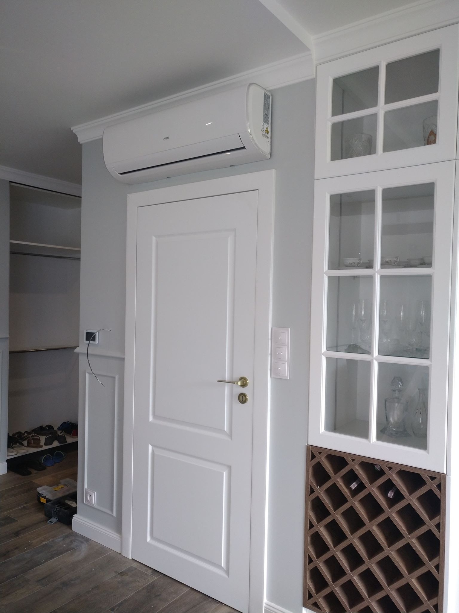 Biała jednostka klimatyzacyjna zamontowana nad białą, dwuskrzydłową wewnętrzną drzwią w pomieszczeniu z białą zabudową meblową z przeszklonymi witrynami i regałem na wino, widoczne narzędzia i buty...