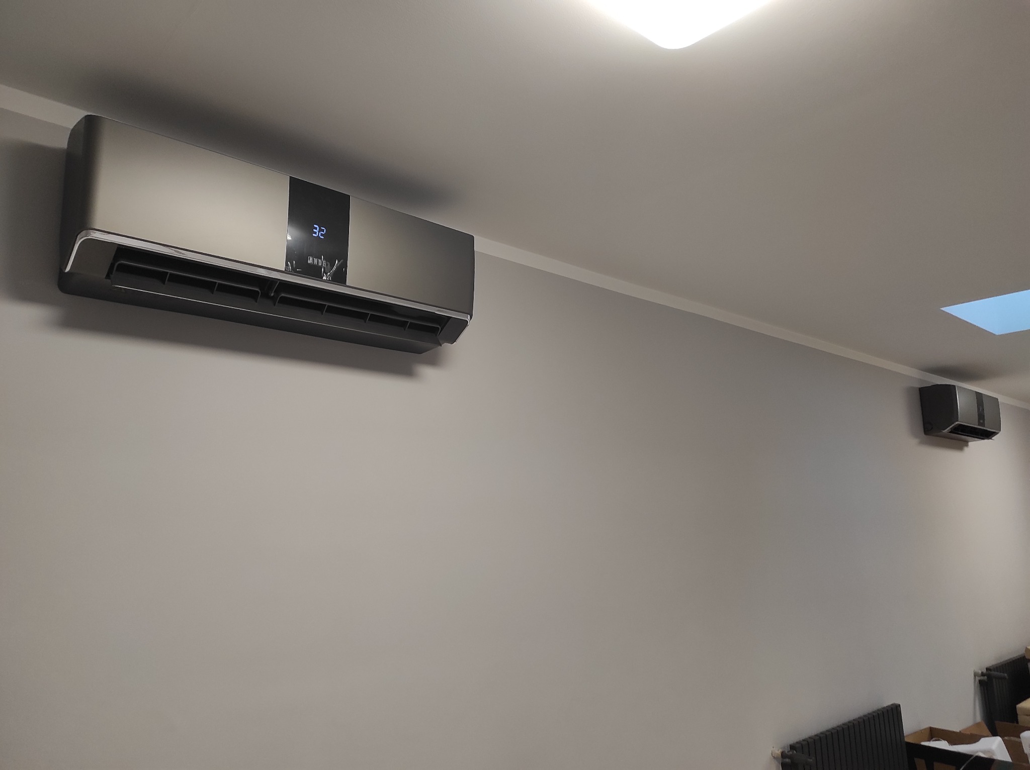 Nowoczesny klimatyzator ścienny w kolorze grafitowym, z wyświetlaczem LED wskazującym temperaturę 32 stopnie, zamontowany na szarej ścianie w pomieszczeniu z grzejnikiem i oknem dachowym.