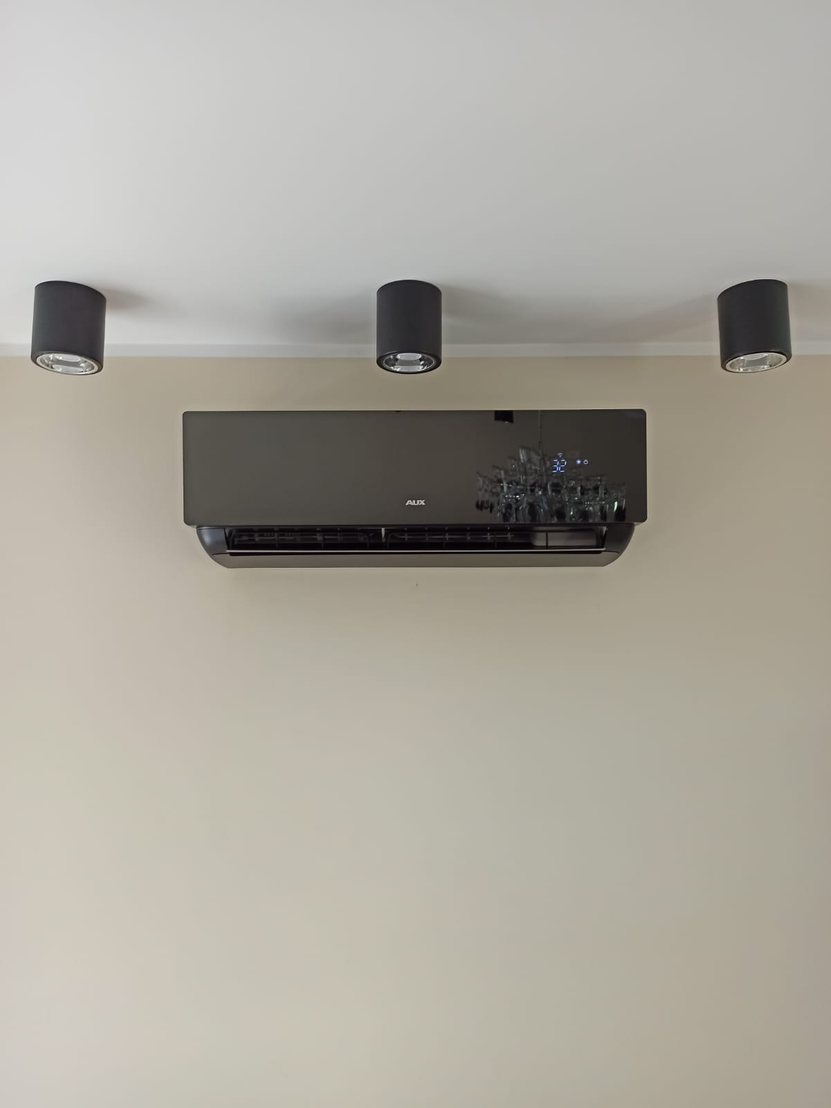 Czarny klimatyzator AUX zamontowany na beżowej ścianie, oświetlony trzema cylindrycznymi lampami sufitowymi.