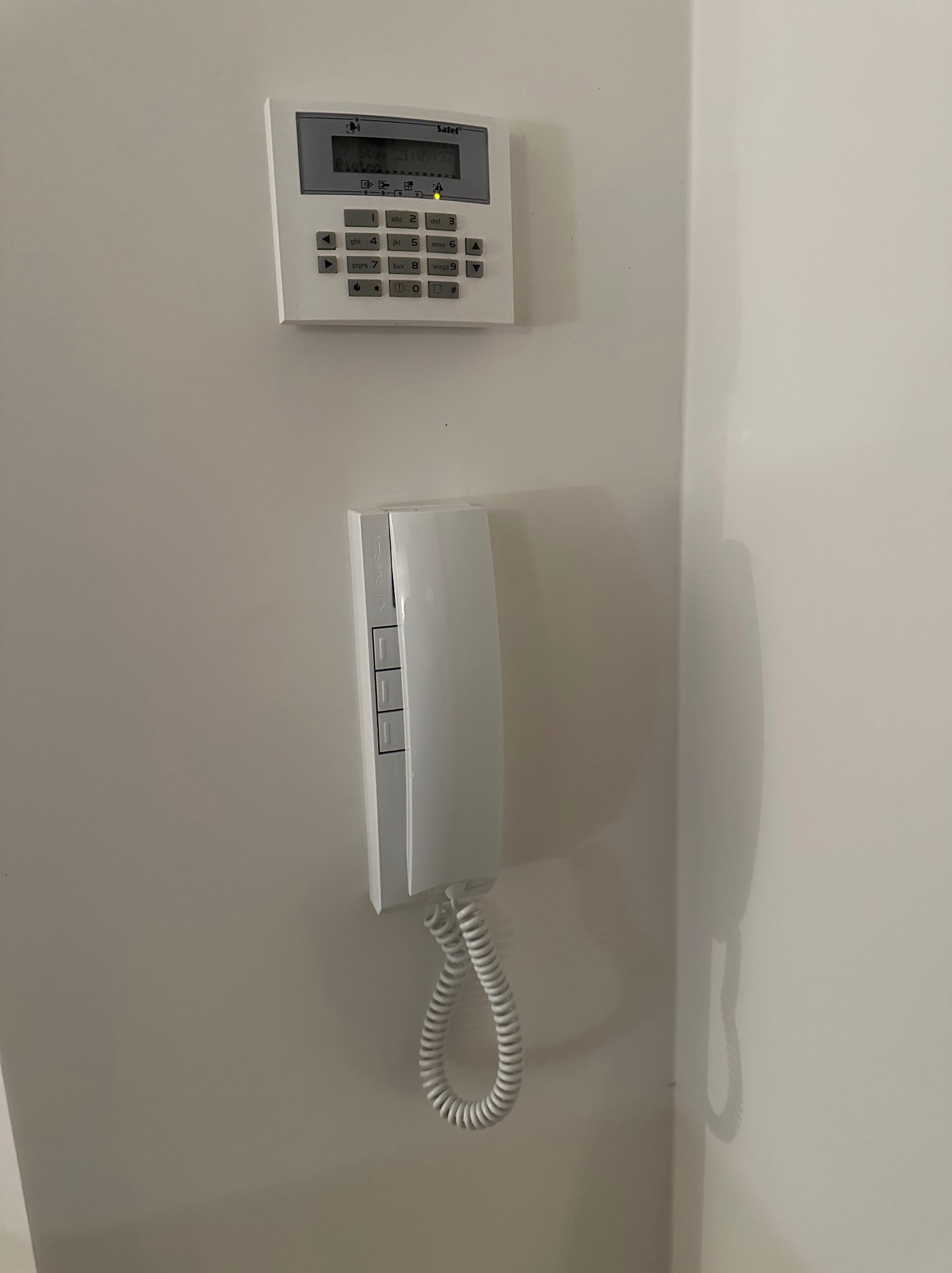 Biały panel alarmowy Safel z klawiaturą i słuchawką domofonu na ścianie. Widoczny wyświetlacz i przyciski. Spiralny kabel słuchawki rzuca cień.