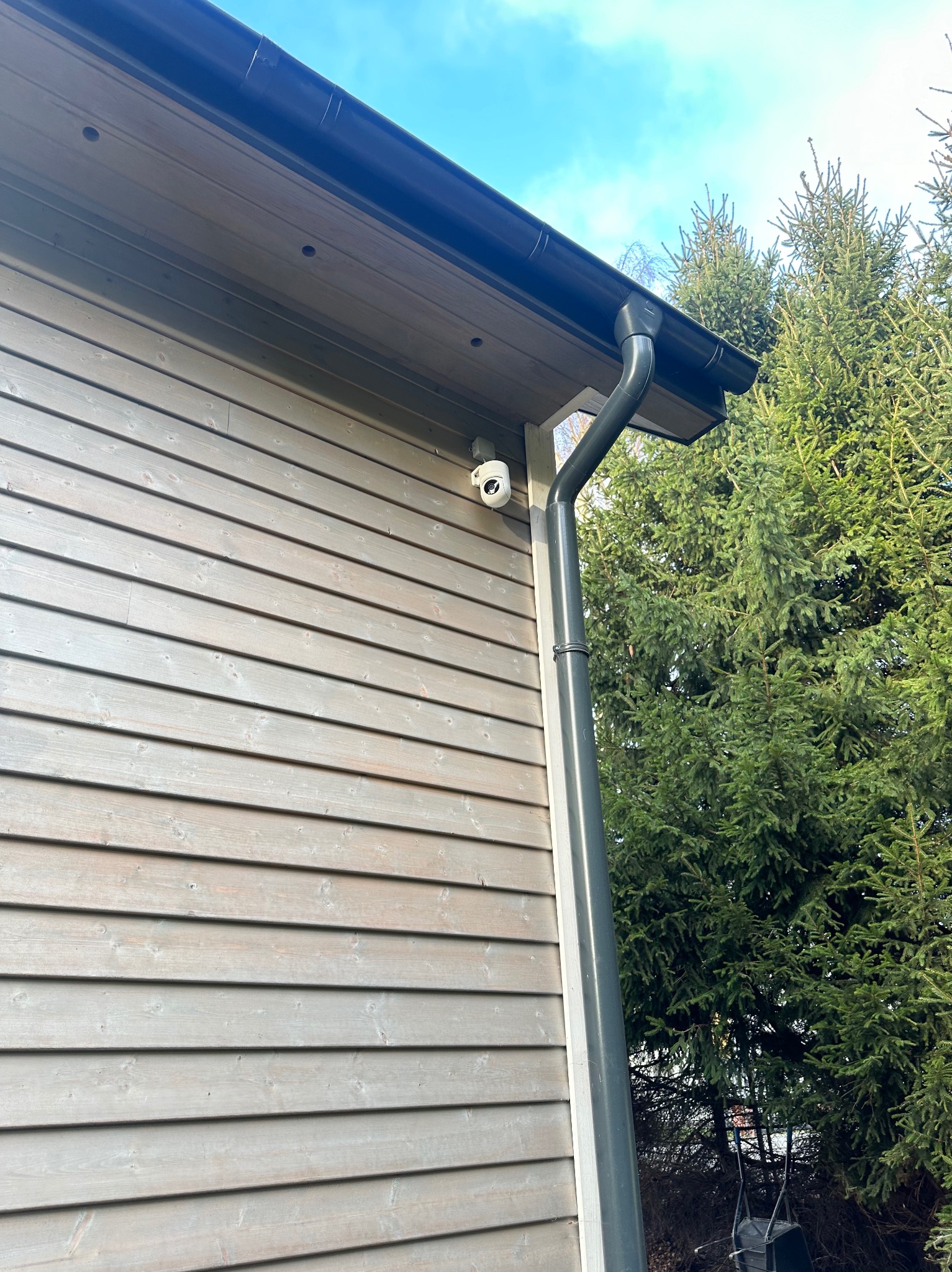 Kamera IP zamontowana na elewacji drewnianego domu, obok rynny spustowej. W tle iglaste drzewa i fragment błękitnego nieba.
