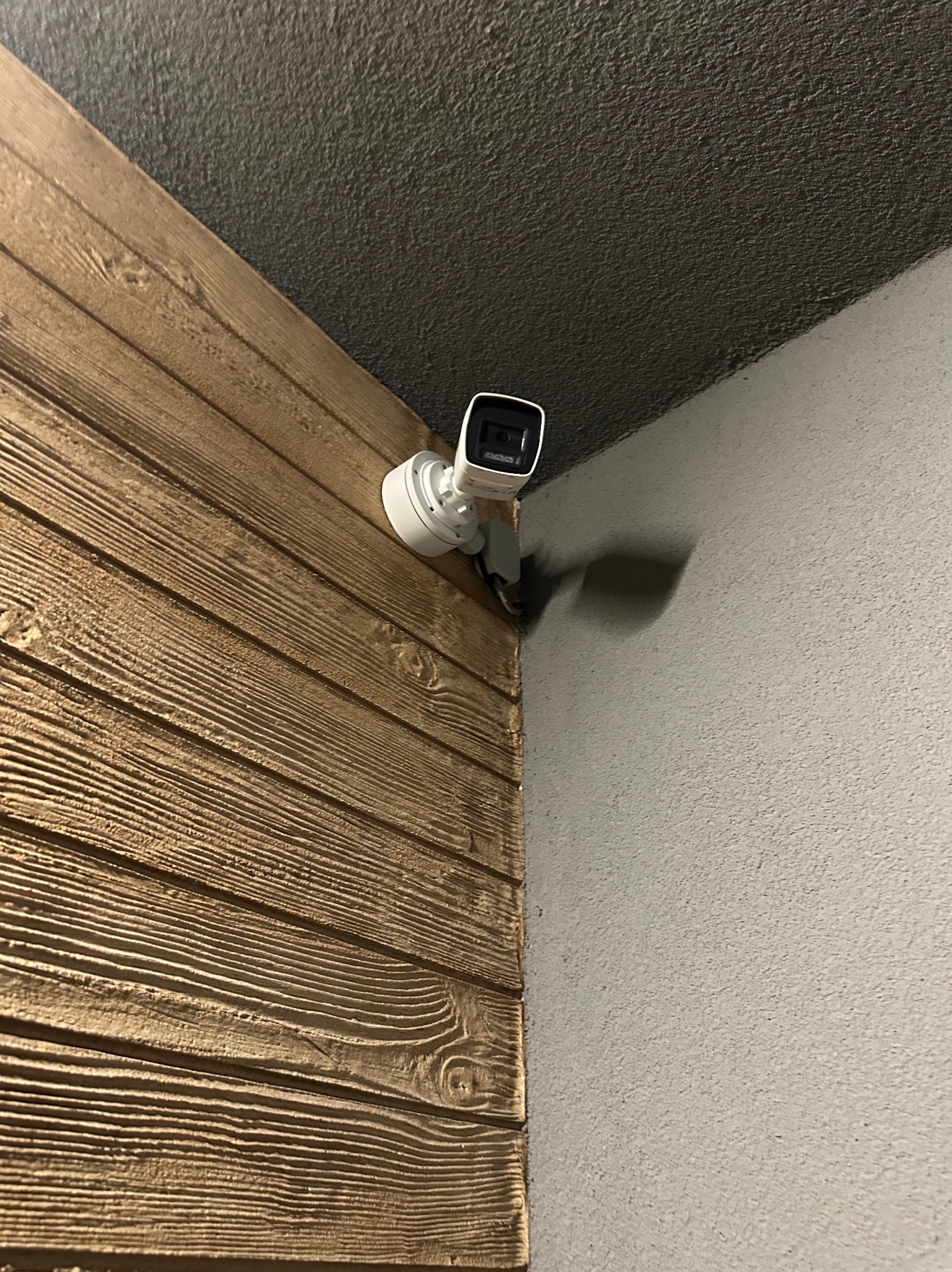 Kamera monitoringu na drewnianej ścianie w Poznaniu. Biała obudowa kamery kontrastuje z teksturą drewna i szarą ścianą. Widok z dołu, podkreślający kąt montażu.