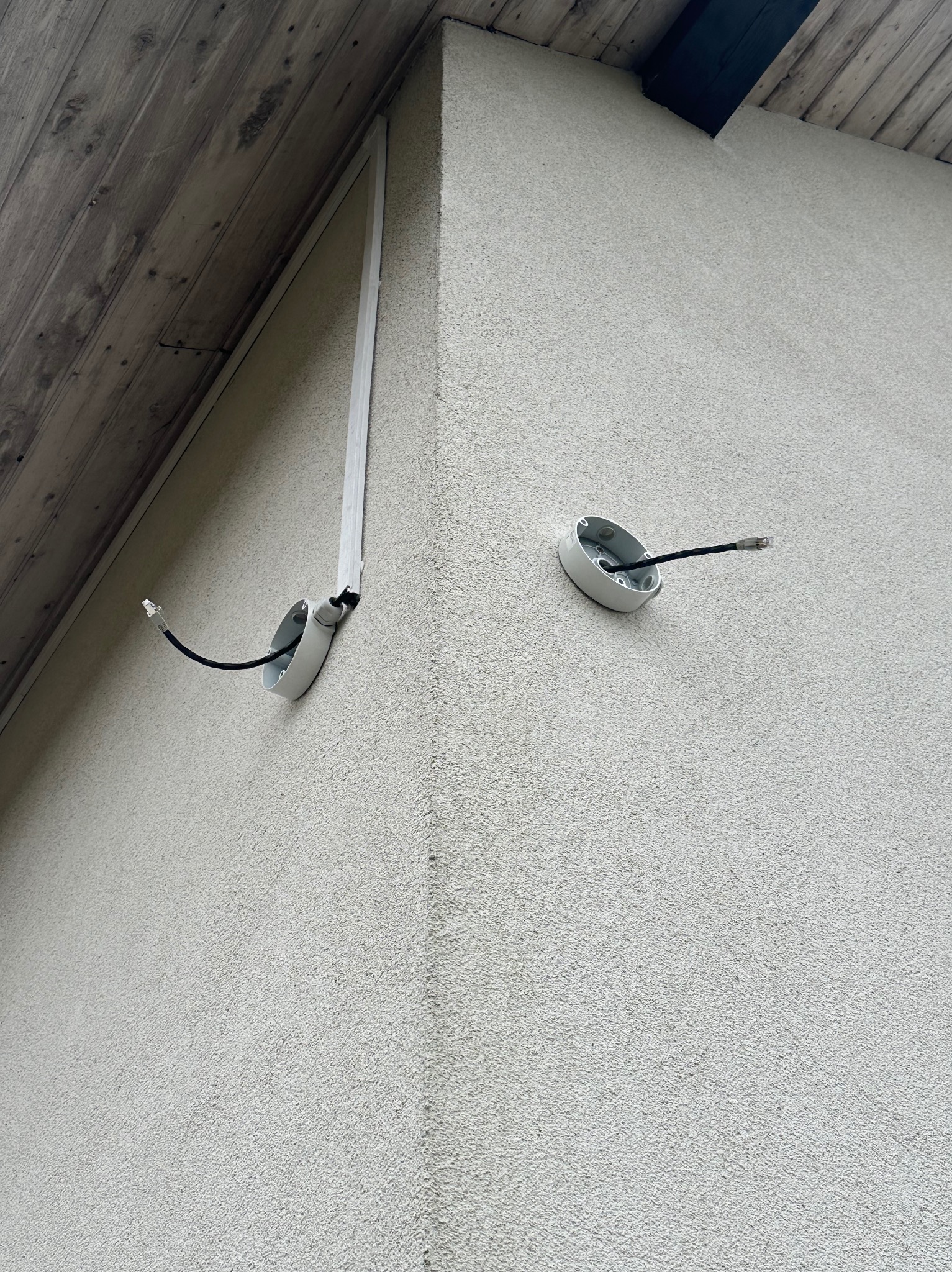 Dwa gniazda z kablami na elewacji budynku, jeden z korytkiem kablowym. Instalacja alarmowa lub monitoringu na zewnątrz. Szara ściana i drewniane elementy dachu.