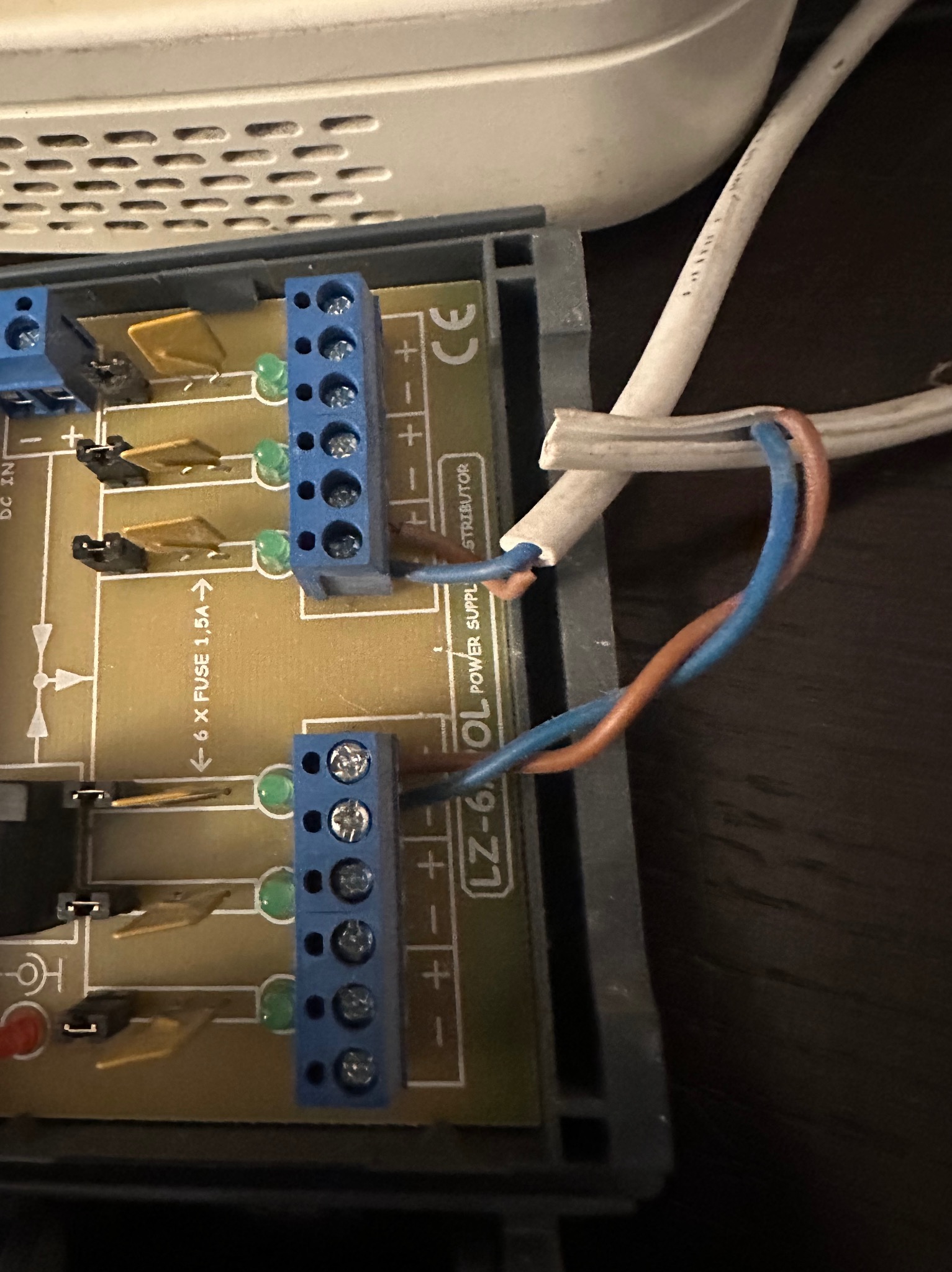 Podłączenie przewodów do listwy zaciskowej wewnątrz obudowy zasilacza. Widoczne bezpieczniki, oznaczenia biegunów i fragment kabla zasilającego.