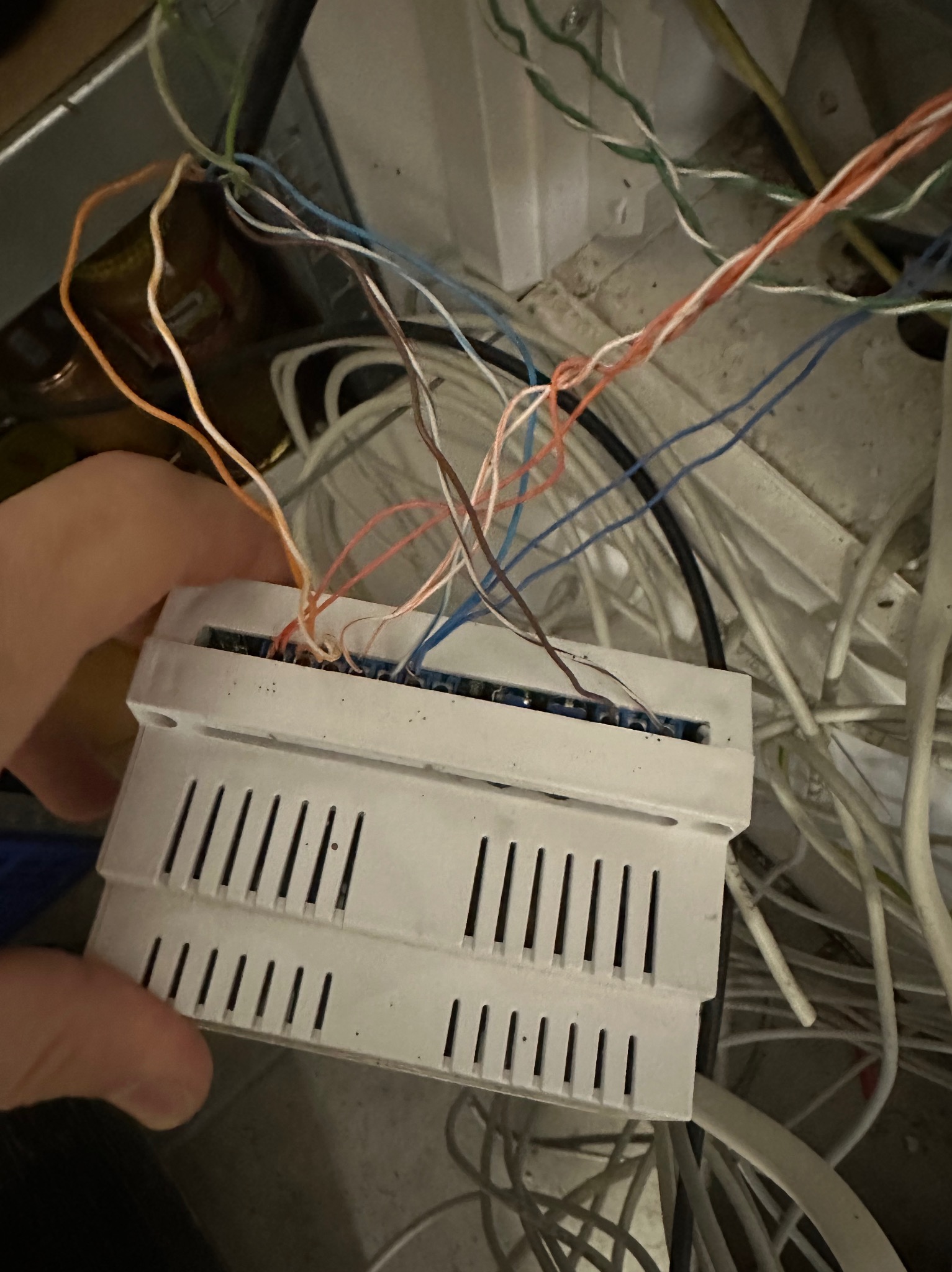 Biała skrzynka z podłączonymi przewodami w dłoni, widoczne liczne kable w tle. Instalacja elektryczna w trakcie montażu lub naprawy.