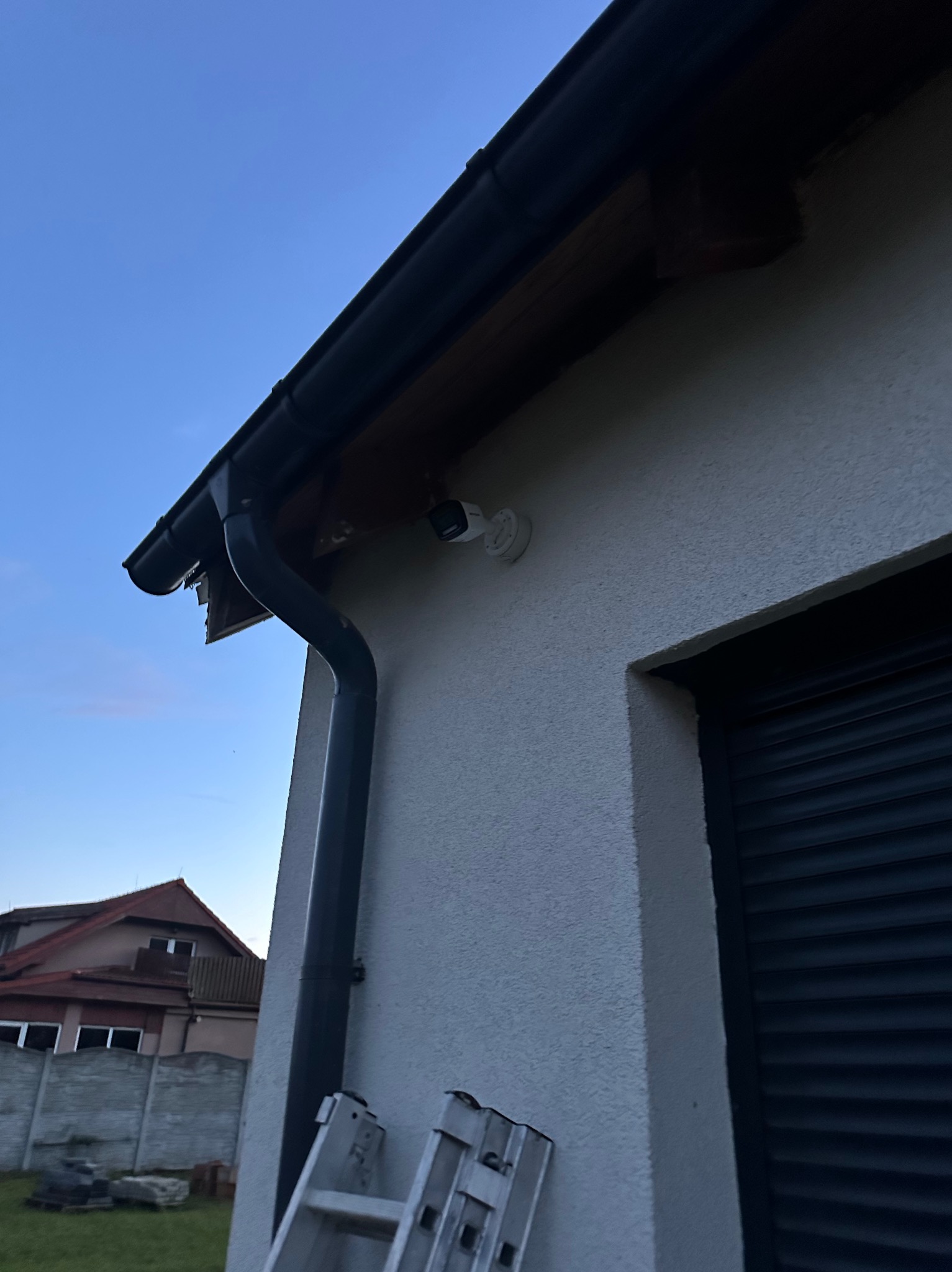Kamera monitoringu zamontowana na elewacji domu, widoczna z dołu, obok rynny i okna z żaluzjami. W tle dom sąsiada i drabina oparta o ścianę.