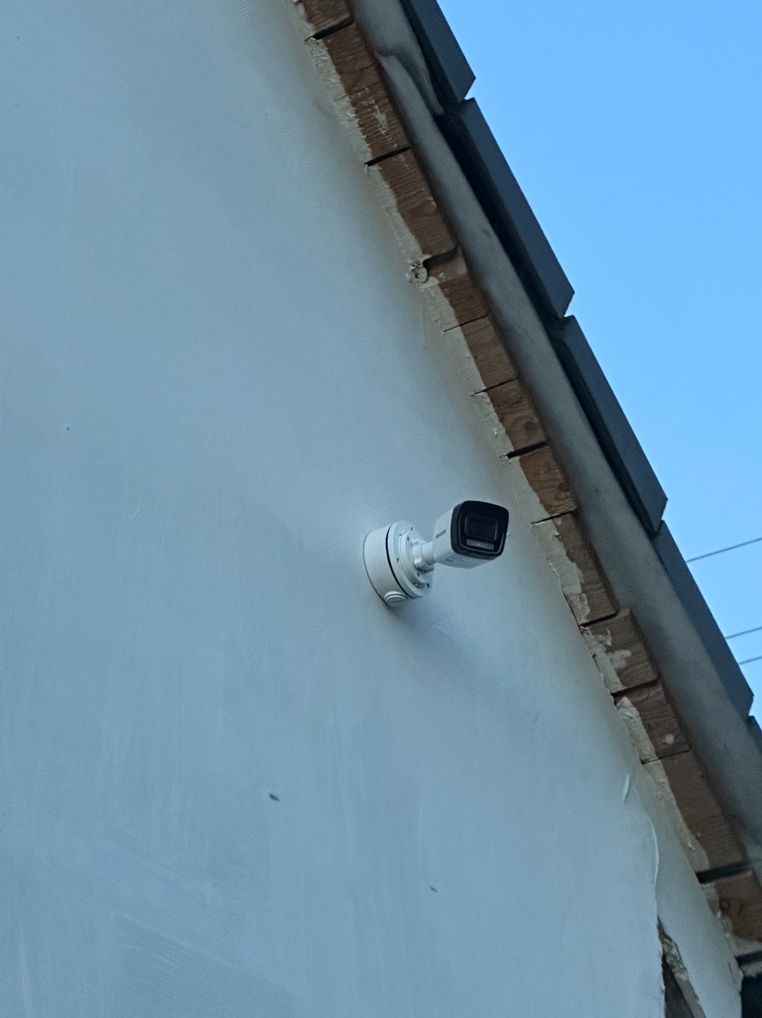 Kamera monitoringu, biała, zamontowana na ścianie budynku z widocznym fragmentem dachu. Ujęcie z dołu, niebo w tle. Widoczny kabel zasilający.